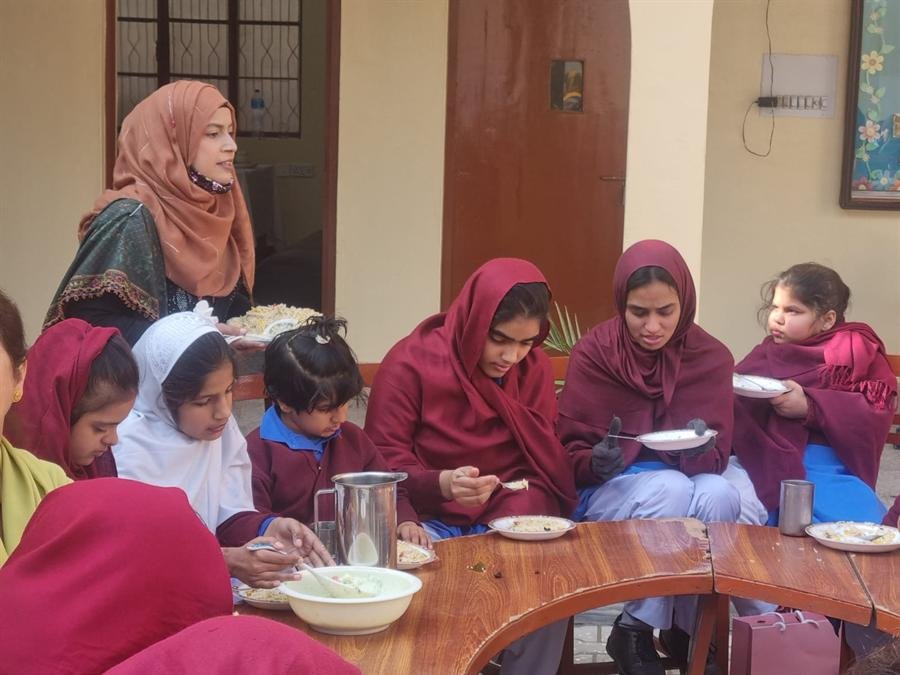 Students in maroon shawls eating together at a round table while a teacher stands nearby