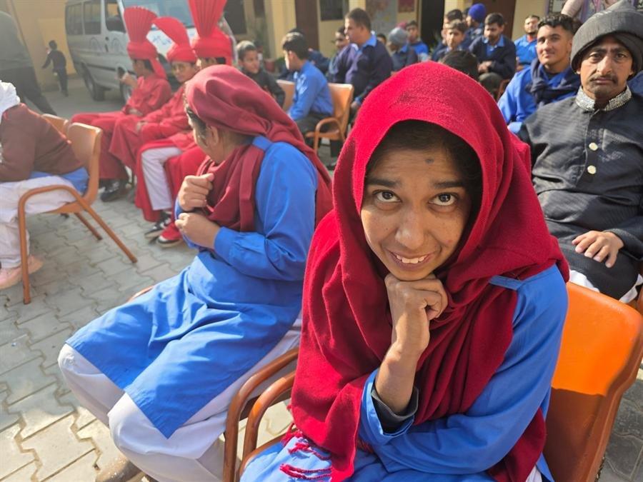 A student in a red shawl smiling with a hand under the chin at an outdoor event